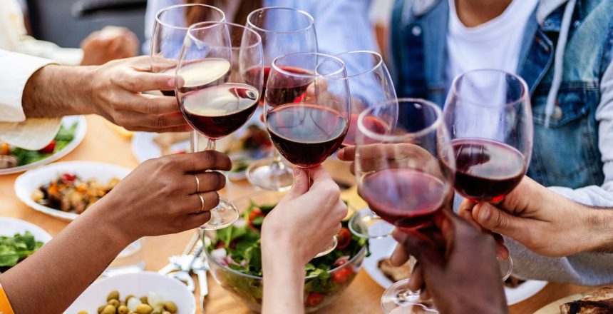 Close-up view of multiracial group of people reunited at summer party toasting red wine together. Outside evening garden celebration with happy friends enjoying dinner barbecue.
